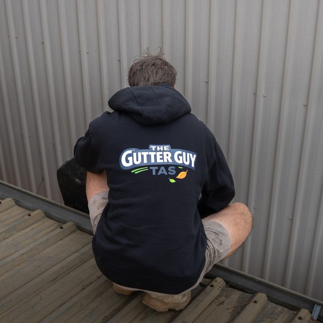 The Gutter Guy Tas crouched on a roof in a branded hoodie, cleaning a gutter